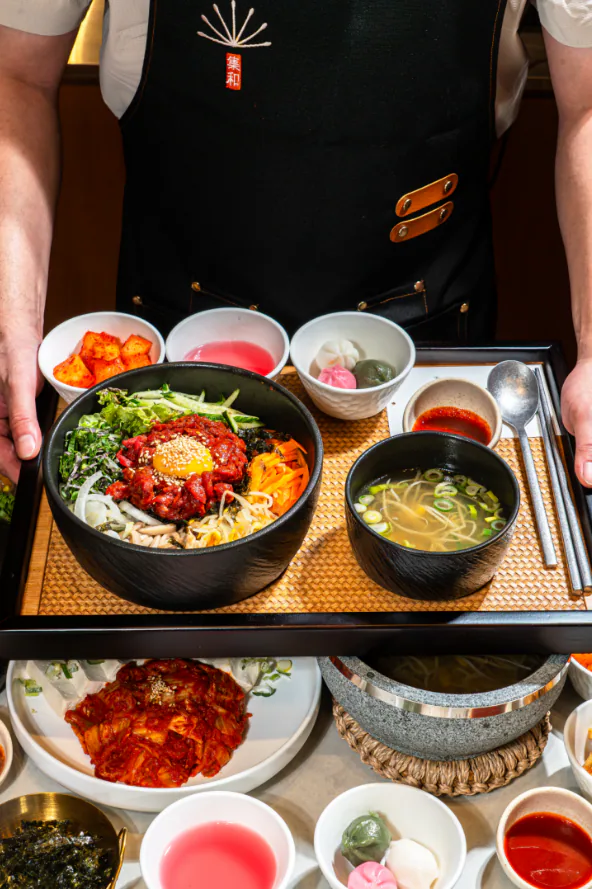 검은 그릇에 담긴 한우육회비빔밥과 국, 반찬이 놓인 라탄 쟁반을 든 직원